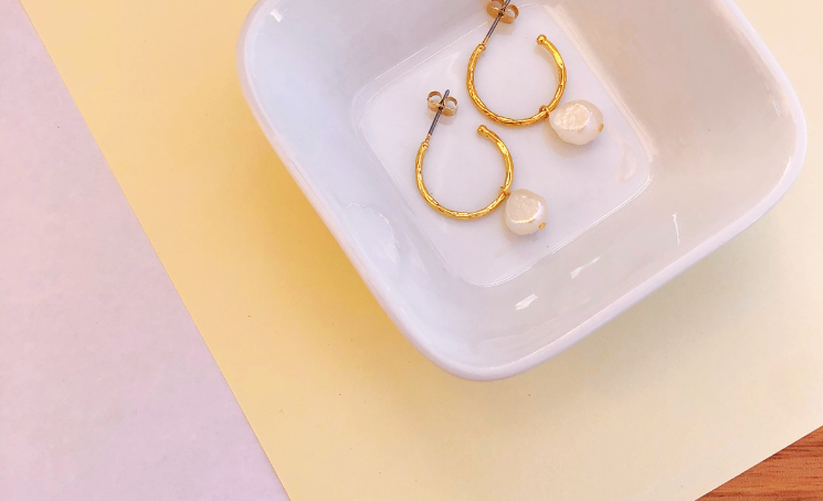 Two gold and pearl huggie hoop earrings in a dish on a white, cream and wood background