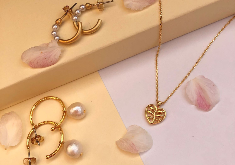 Two pairs of Joya Jewellery gold huggie earrings with pearls and a gold pendant necklace with a heart leaf designed pendant with flower petals scattered over jewellery