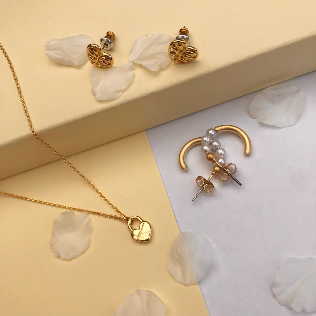 Gold heart shaped studs on a step with a pair of gold Huggies with pearls below and a gold necklace with a heart lock shaped pendant.